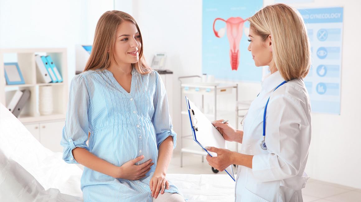 Młoda, uśmiechnięta kobieta w ciąży siedzi na kozetce i trzyma się za brzuch podczas rozmowy z lekarką.