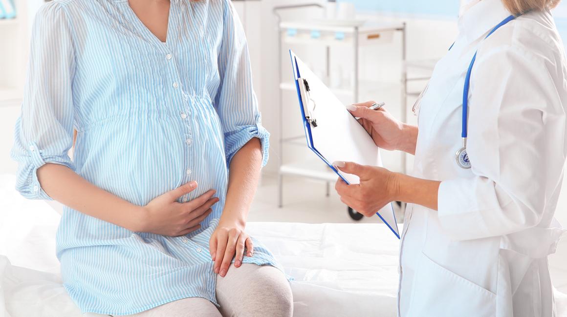 Młoda, uśmiechnięta kobieta w ciąży siedzi na kozetce i trzyma się za brzuch podczas rozmowy z lekarką.