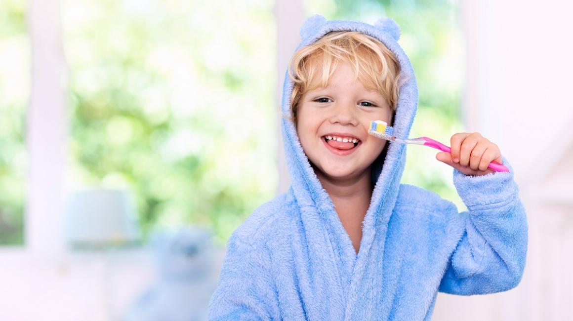 Kilkuletni chłopczyk w szlafroku trzyma szczoteczkę do zębów. Wystawia język, widać mu zęby mleczne.