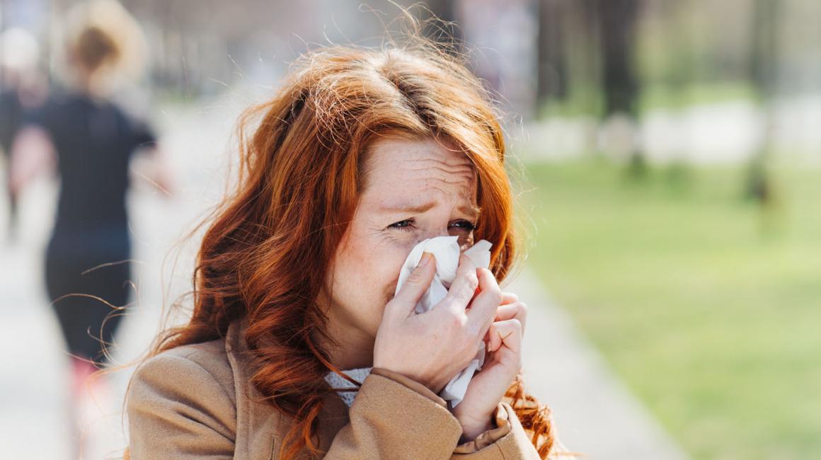 Kobieta, która jest uczulona na pyłki, kicha i zasłania nos chusteczką. Stoi na chodniku, ma rude włosy.