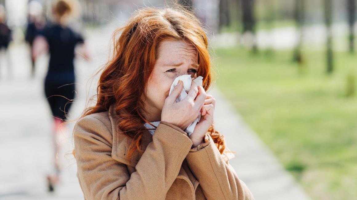 Kobieta, która jest uczulona na pyłki, kicha i zasłania nos chusteczką. Stoi na chodniku, ma rude włosy.