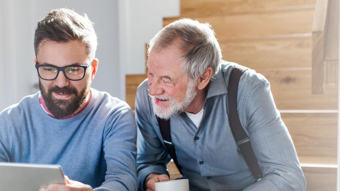 Dorosły syn z ojcem siedzą na schodach w domu. Syn pokazuje coś ojcu na tablecie, senior ma w dłoniach kubek.