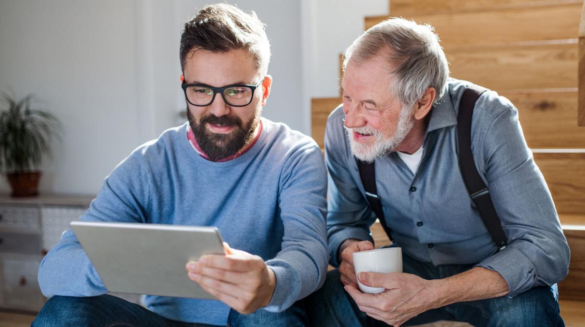 Dorosły syn z ojcem siedzą na schodach w domu. Syn pokazuje coś ojcu na tablecie, senior ma w dłoniach kubek.