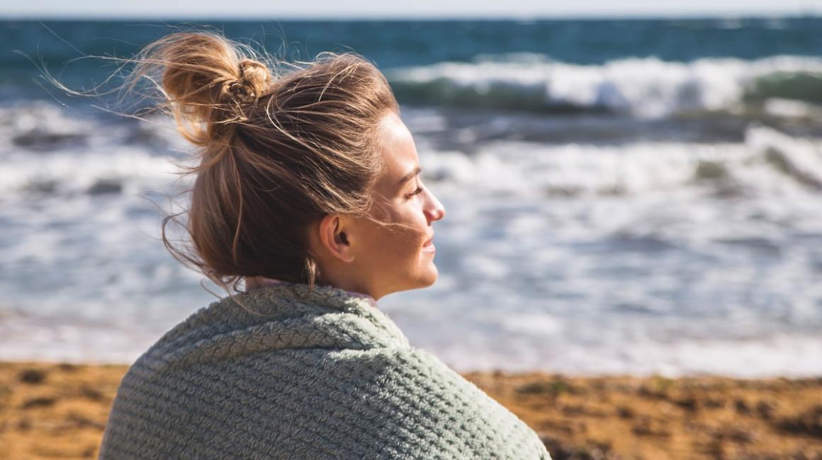 Młoda kobieta w blod włosach spiętych w koczek siedzi na plaży. Patrzy w stronę morza, słońce oświetla jaj twarz. Na ramionach ma koc.