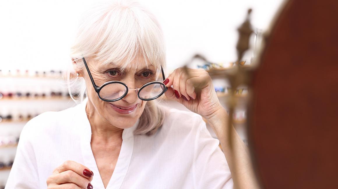 Starsza pani przymierza okulary u optyka. Ma siwe włosy i czerwone paznokcie.
