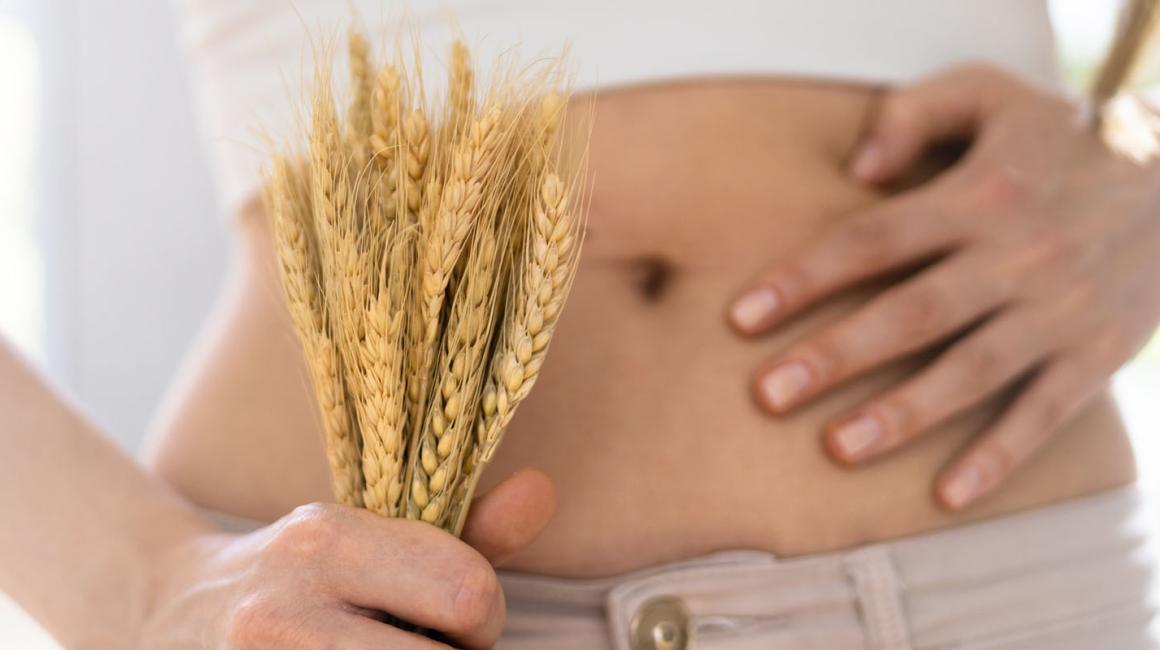 Zbliżenie na goły brzuch kobiety. W jednej ręce trzyma kłosy zbóż, a drugą dotyka brzucha.