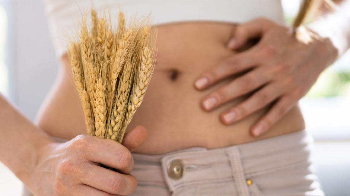 Zbliżenie na goły brzuch kobiety. W jednej ręce trzyma kłosy zbóż, a drugą dotyka brzucha.