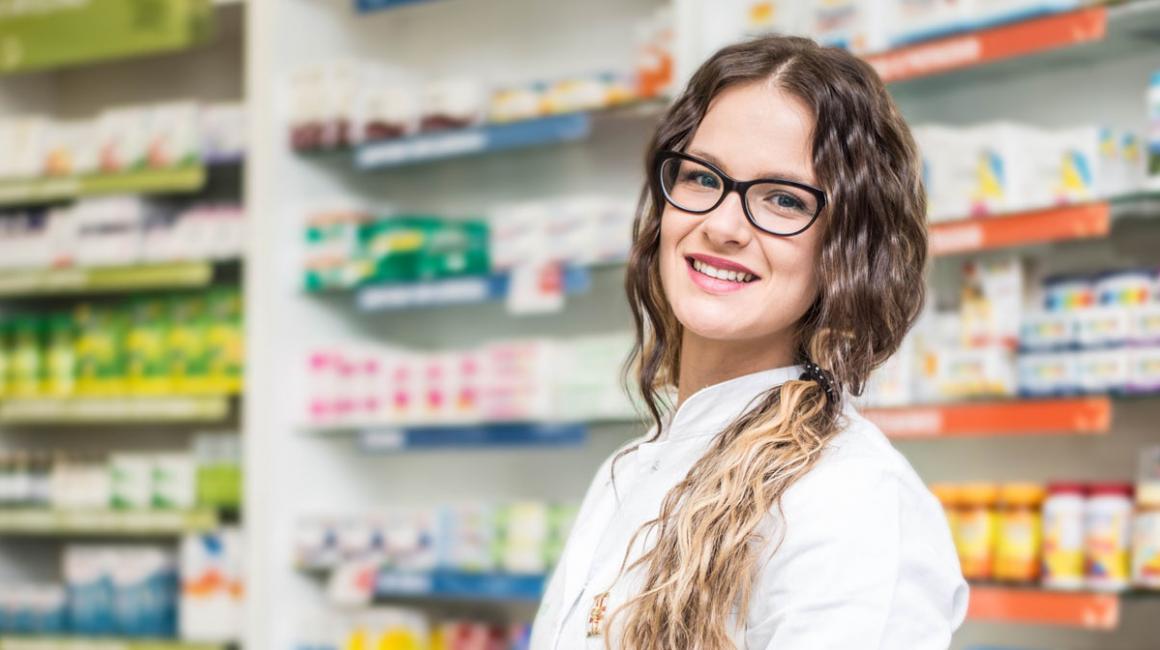 Uśmiechnięta farmaceutka w aptece. Młoda dziewczyna ma długie, falowane włosy upięte w kucyk na boku. Ma okulary. 