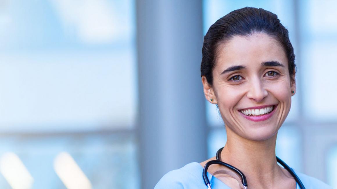 Uśmiechnięte pielęgniarka stoi przed przychodnią. Na szyi ma stetoskop, w tle stoi lekarz.