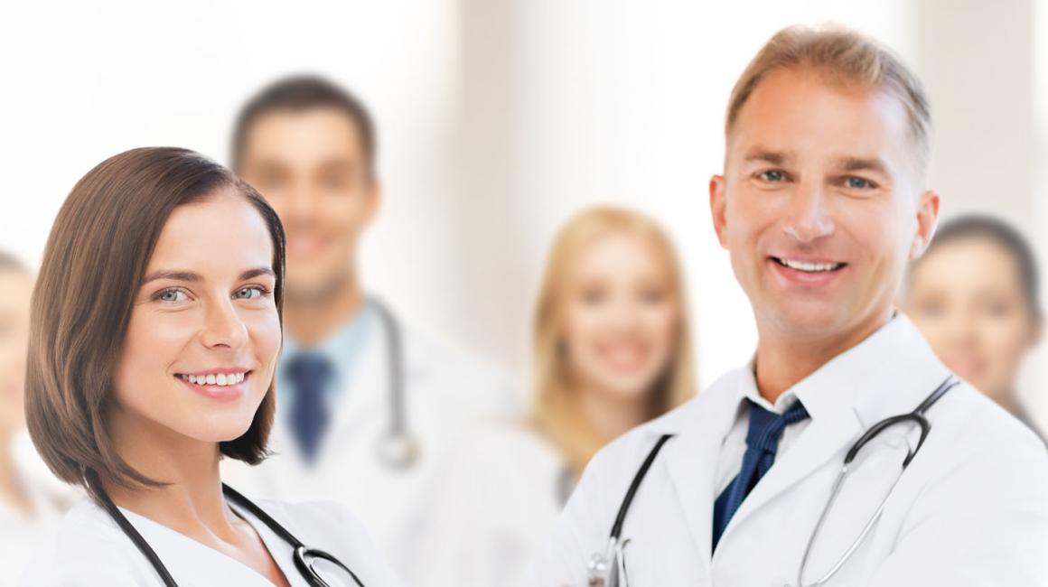 Na zdjęciu jest grupa usmiechniętych lekarzy w kitlach. Po lewej stronie stoi kobieta, po prawej mężczyzna, reszta niewyraźna w tle.