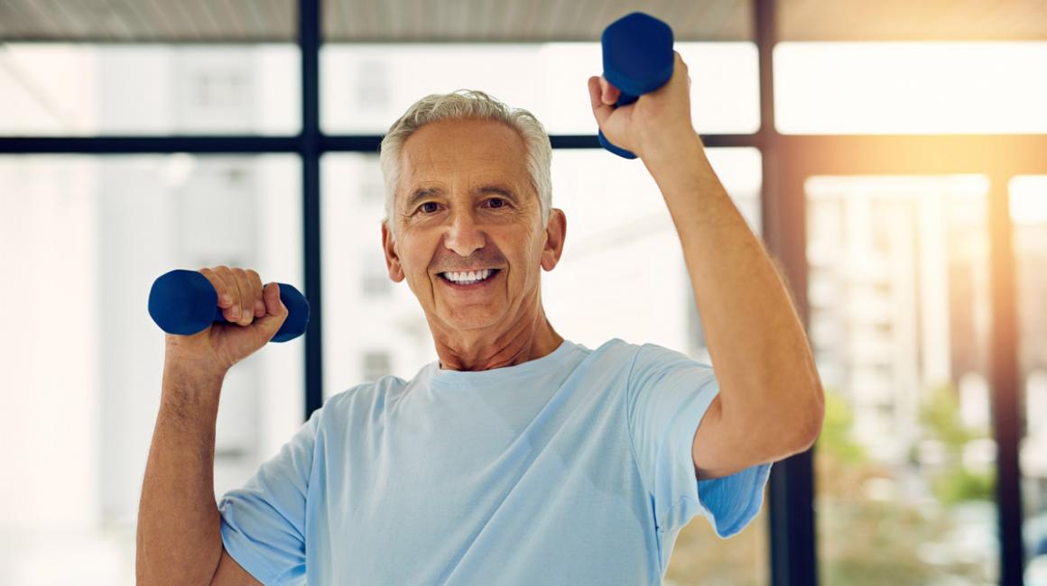 Starszy mężczyzna ćwiczy. Podnosi ciężarki na wysokość głowy. Ma siwe włosy, błękitny t-shirt. Uśmiecha się.