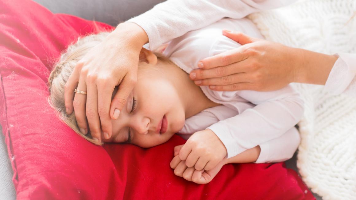 Matka trzyma dłoń na czole córki, sprawdza jej tempreraturę. Dziewczynka leży pod kocem. 