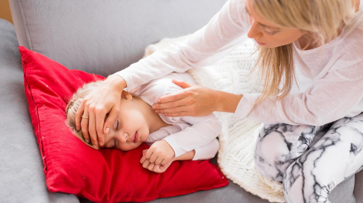 Matka trzyma dłoń na czole córki, sprawdza jej tempreraturę. Dziewczynka leży pod kocem.