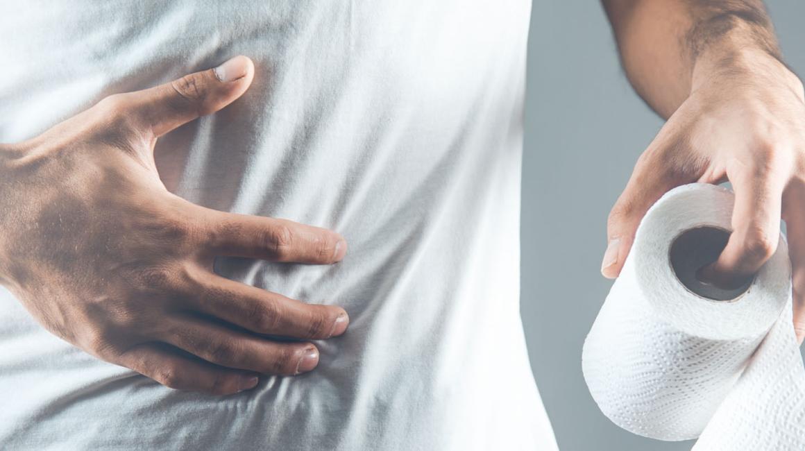 Mężczyzna w białej koszulce. Zbliżenie na jego bolący brzuch, na którym trzyma rękę. W drugiej ma rolkę papieur toaletowego.