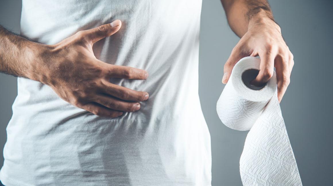 Mężczyzna w białej koszulce. Zbliżenie na jego bolący brzuch, na którym trzyma rękę. W drugiej ma rolkę papieur toaletowego.