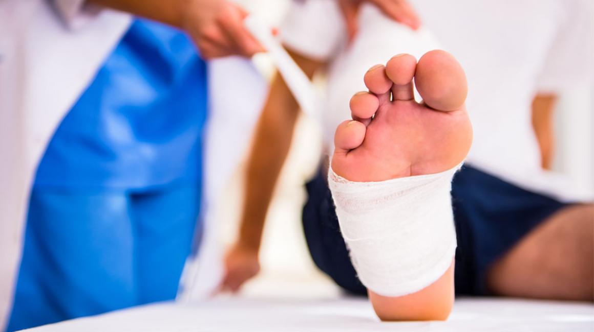 Mężczyzna podczas wizyty u ortopedy. Na zbliżeniu widać jego stopę owiniętą bandażem.