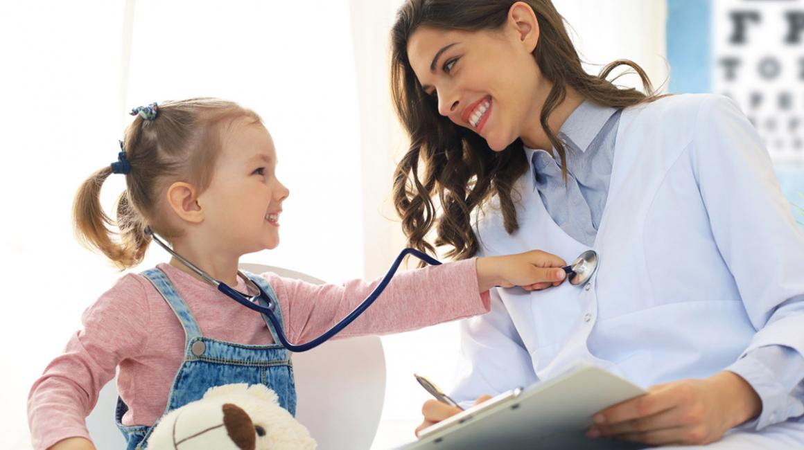 Mała dziewczynka podczas wizyty kontrolnej u lekarza. Na kolanach ma misia, na szyi stetoskop, którym bada lekarkę. Obie się uśmiechają