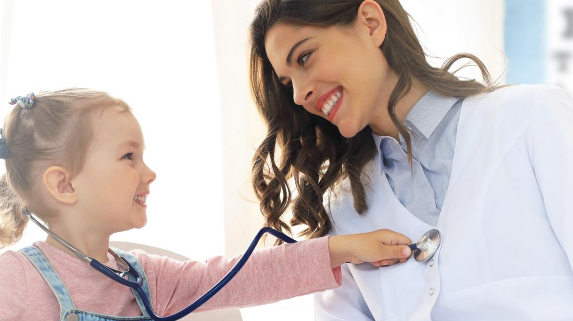 Mała dziewczynka podczas wizyty kontrolnej u lekarza. Na kolanach ma misia, na szyi stetoskop, którym bada lekarkę. Obie się uśmiechają