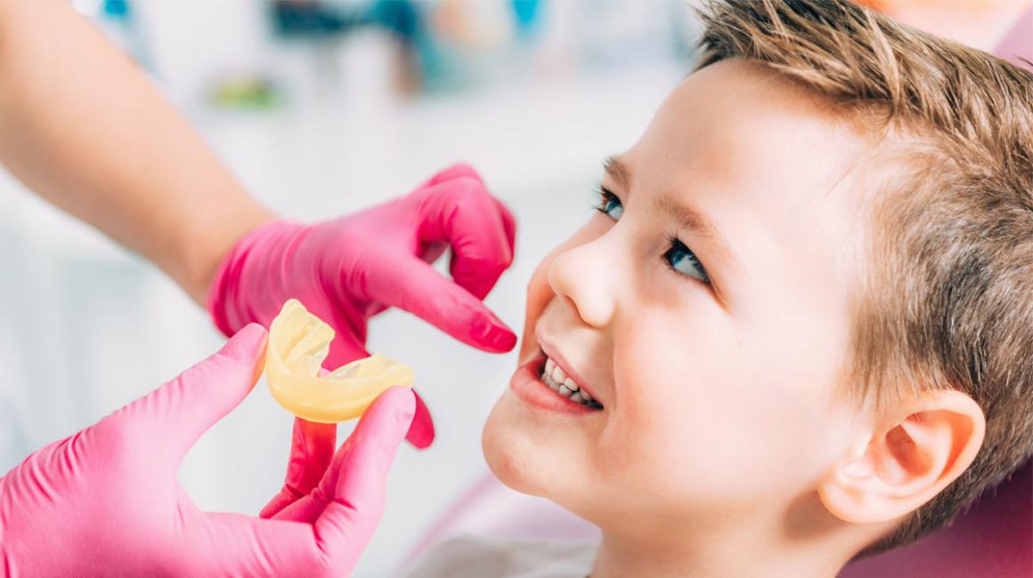 Chłopiec podczas wizyty u ortodonty. Lekarz w różowych rękawiczkach zbliża wycisk do jego ust.