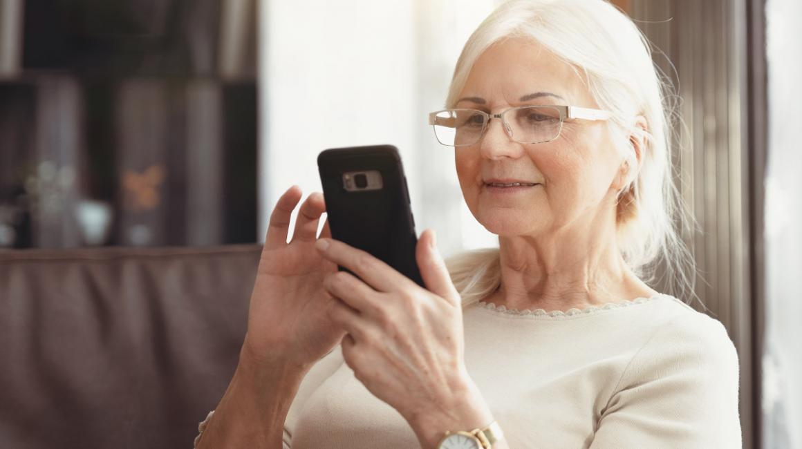 Seniorka w domu siedzi na sofie, ma w ręce telefon i coś sprawdza. Ma siwe, długie włosy i okulary.