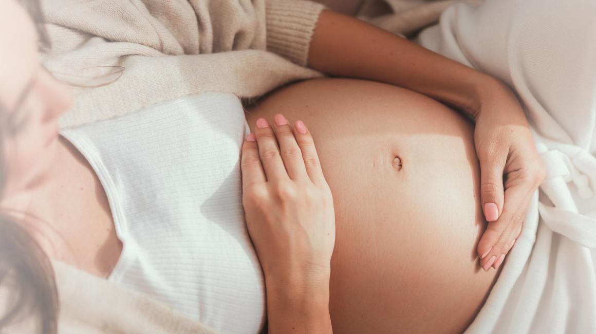 Leżąca kobieta w ciąży obejmuje brzuch dłońmi.