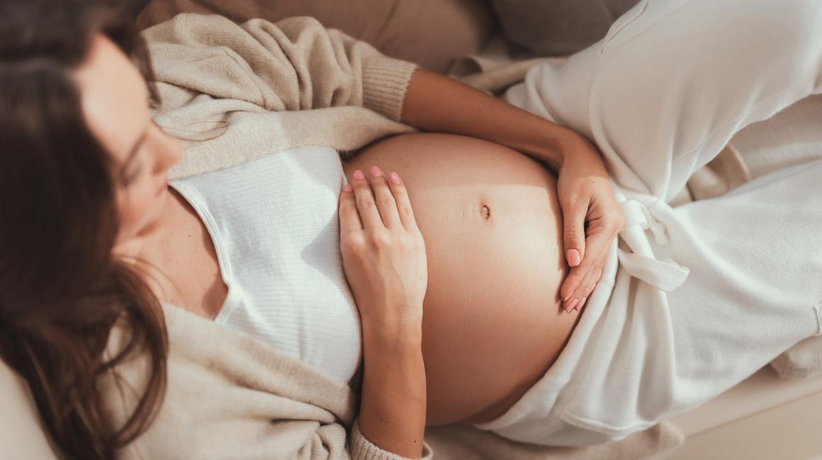 Leżąca kobieta w ciąży obejmuje brzuch dłońmi.