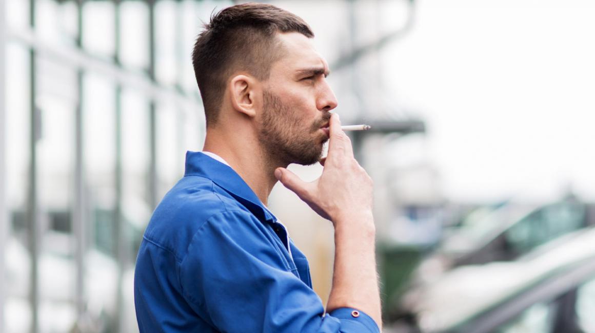 Profil mężczyzny, który stoi przed warsztatem i pali papierosa. Jest ubrany w kombinezon, ma lekki zarost.