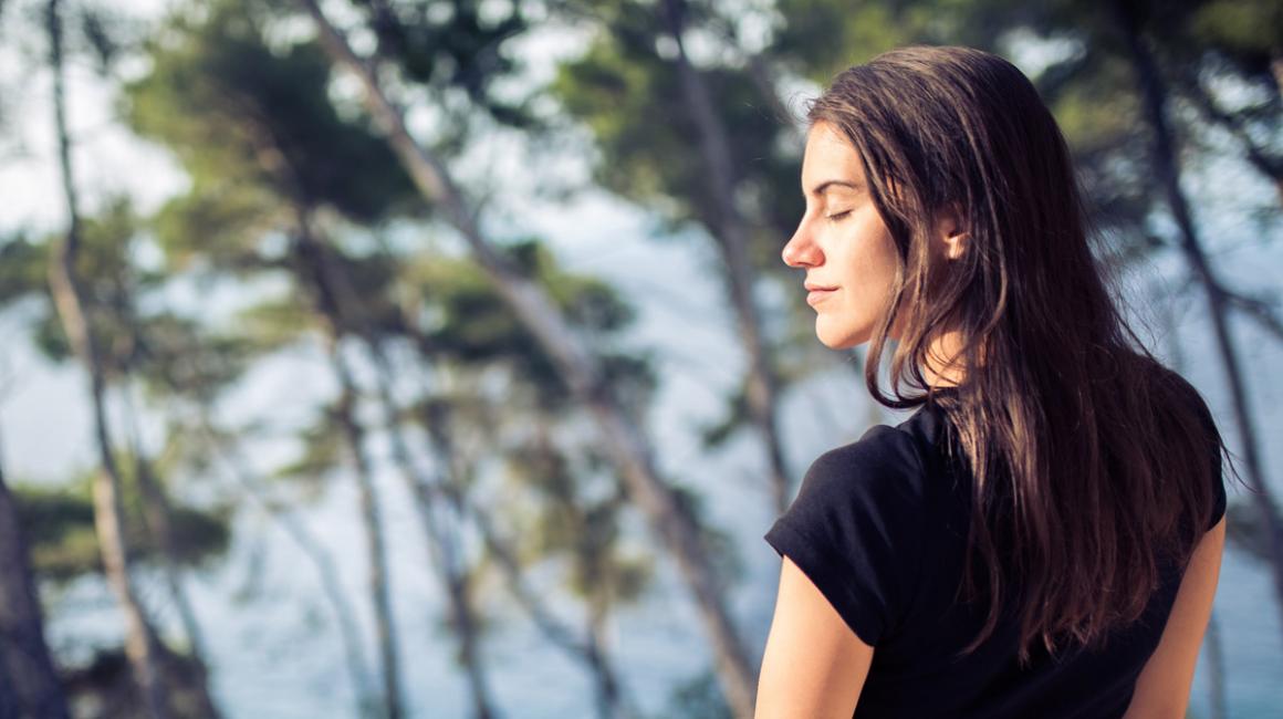 Kobieta stoi nad morzem, w tle rosną sosny. Ma zamknięte oczy, twarz wystawia do słońca. Delektuje się chwilą