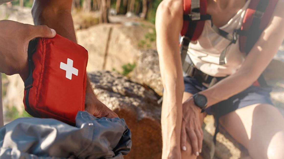 Dziewczyna trzymająca się za kolano, z grymasem bólu. Obok osoba z apteczką w ręce.