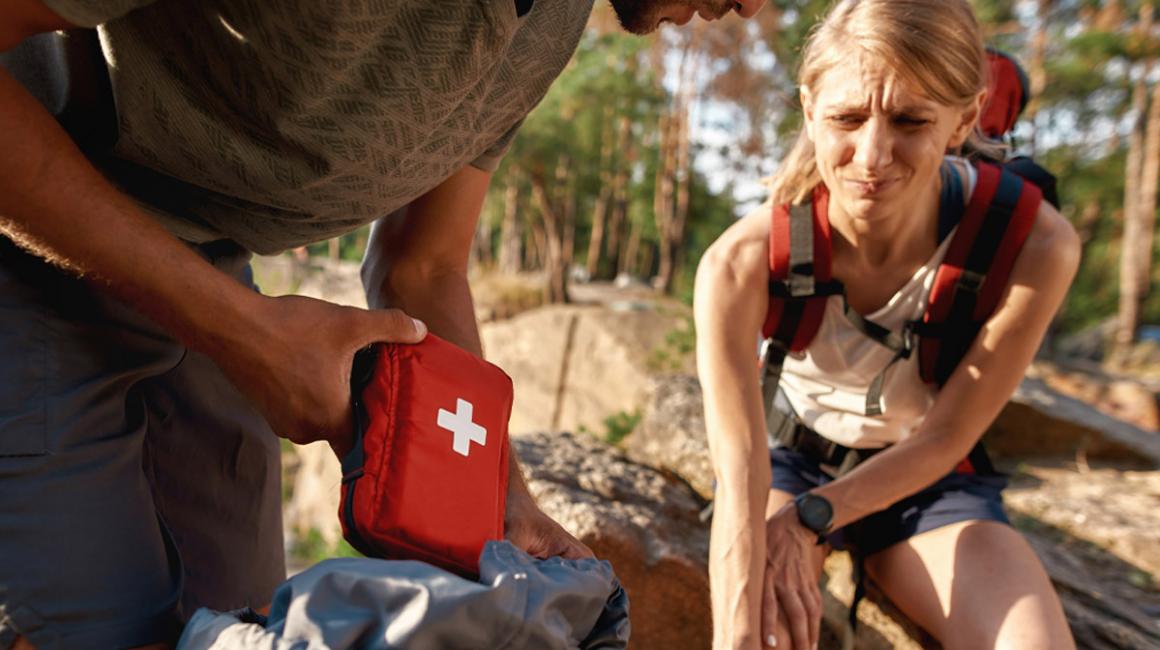 Dziewczyna trzymająca się za kolano, z grymasem bólu. Obok osoba z apteczką w ręce.