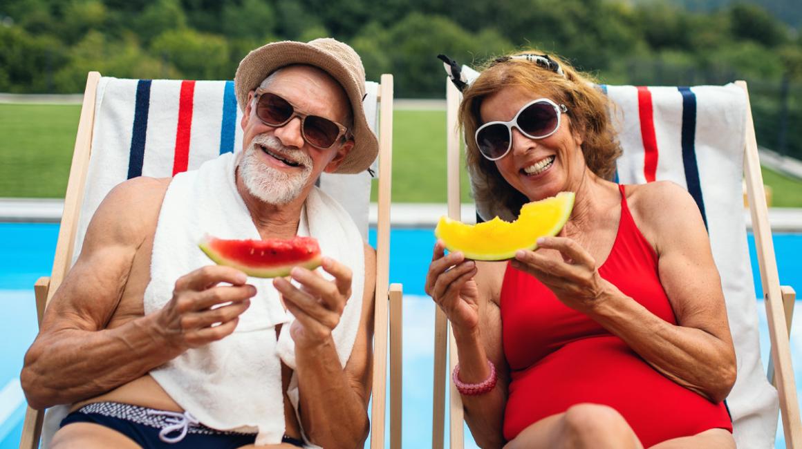 Mężczyzna i kobieta na leżakach, nad basenem. W strojach kąpielowych i okularach przeciwsłownecznych. Jedzą arbuzy. 