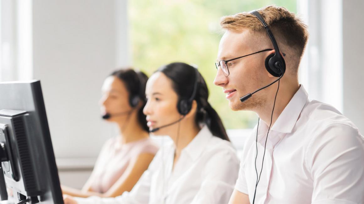 3 konsultantów w słuchawkach na uszach, siedzą przed ekranami komputera. Ujęcie z profilu, od pasa w górę. 