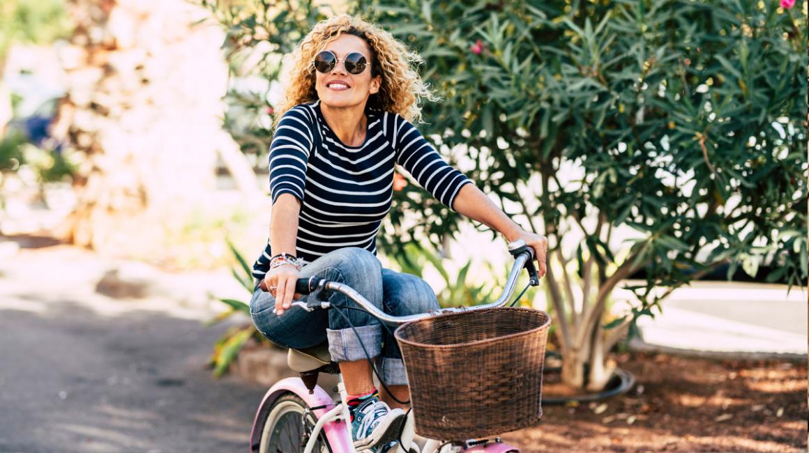 Kobieta w okularach przeciwsłonecznych jadąca na rowerze z wiklinowym koszykiem z przodu. Nogi trzyma na ramie.