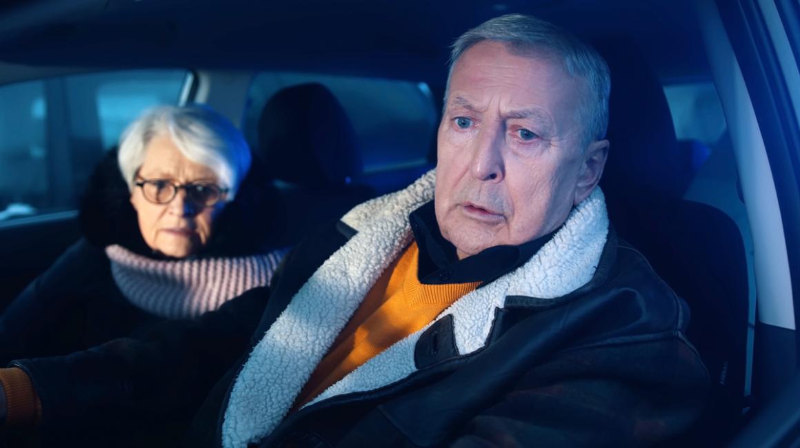 Starsza para w aucie nocą. Mężczyzna za kierownicą jest zmieszany, kobieta lekko wystraszona.