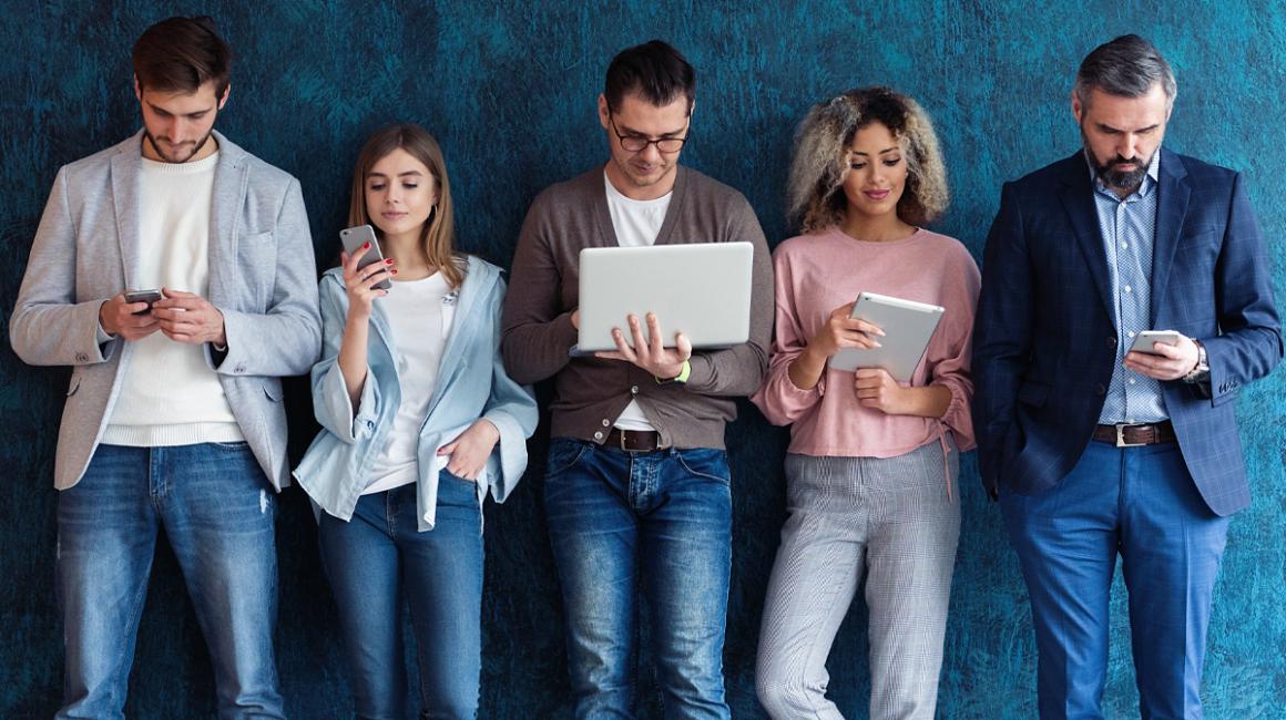 Piatka różnych ludzi stoi przed niebieską ścianą. Trzymają różne urządzenia elektroniczne.