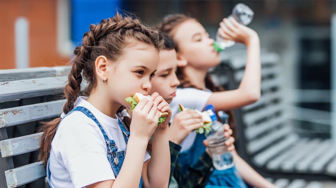 Trójka dzieci siedzi na ławce przed szkołą i je drugie śniadanie. Na pierwszym planie dziewczyna z kanapką z której wystaje sałata, w tle inna pije wodę.