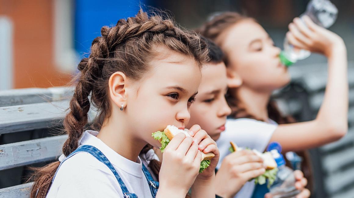 Trójka dzieci siedzi na ławce przed szkołą i je drugie śniadanie. Na pierwszym planie dziewczyna z kanapką z której wystaje sałata, w tle inna pije wodę.