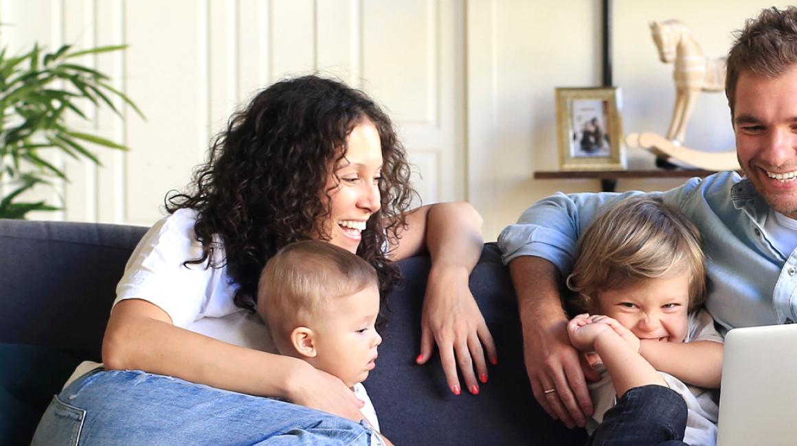 Czteroosobowa rodzina na kanapie w domu. Patrzą na laptop, który ojciec trzyma.