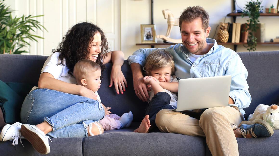 Czteroosobowa rodzina na kanapie w domu. Patrzą na laptop, który ojciec trzyma.