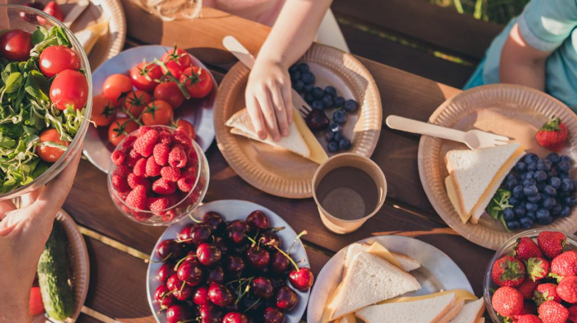 Widok z góry na stoł, na którym są na talerzach pomidory, maliny, czereśnie, truskawki, kanapki z serem oraz roszponka.