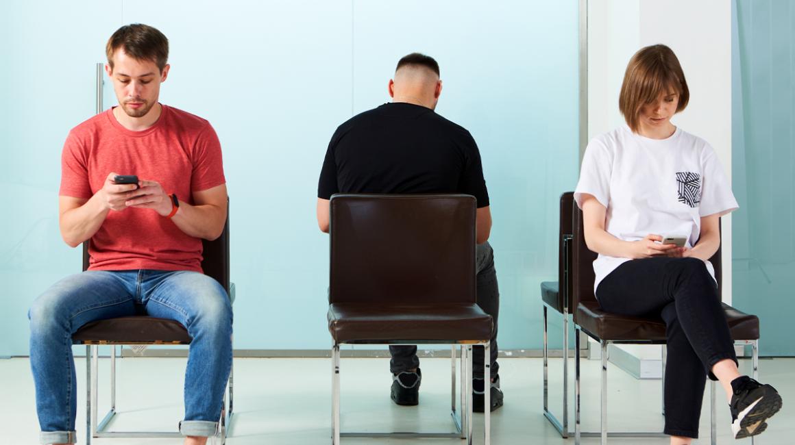 Trzy osoby siedzą na krzesłach w poczekalni w przychodni. Patrzą w telefony.