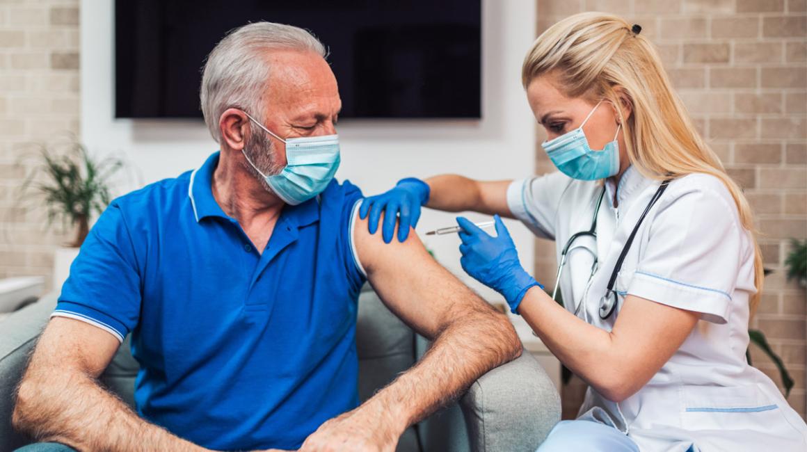 Straszy mężczyna w maseczce siedzący na fotelu. Młoda bobieta w białym fartuchu podaje mu szczepionkę w ramię.
