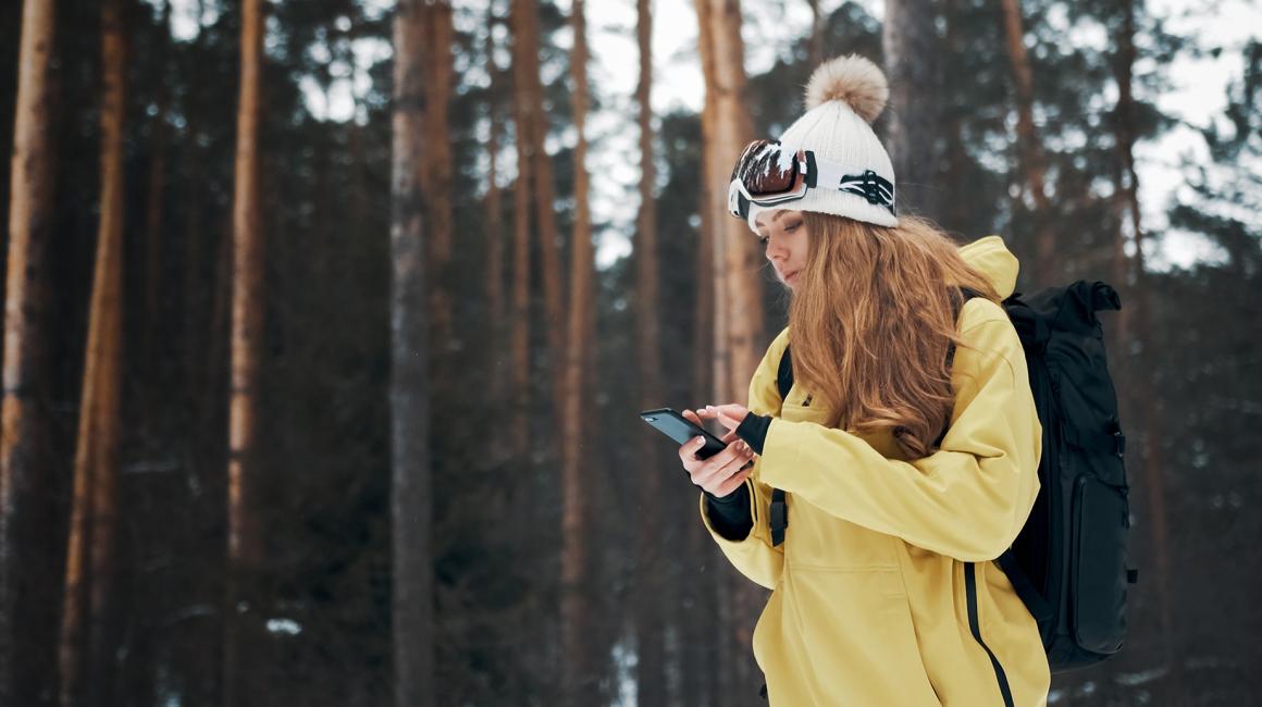 Dziewczyna w żółtej kurtce i czapce na tle lasu. Sprawdza coś w telefonie.