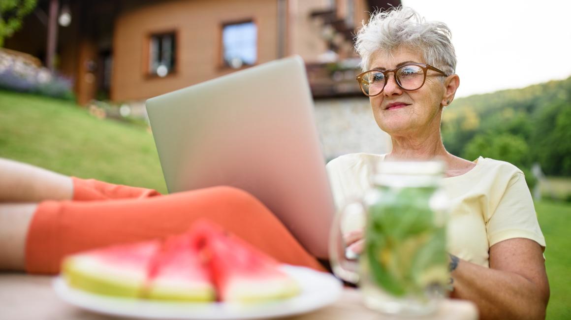Siwa pani z laptopem przed domem w górach, laptop na kolanach, nogi na stole, na stole arbuz, w tle dom na stoku