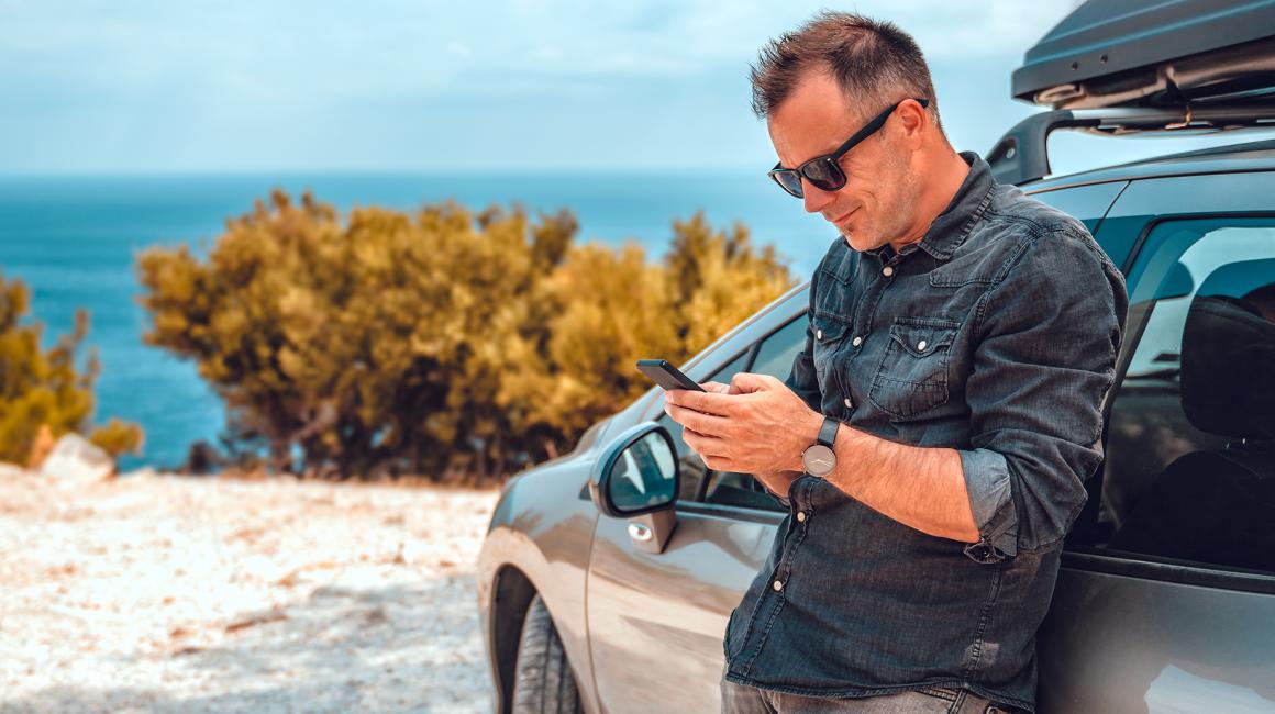 Mężczyzna oparty o auto, z telefonem, w tle krajobraz środziemnomorski