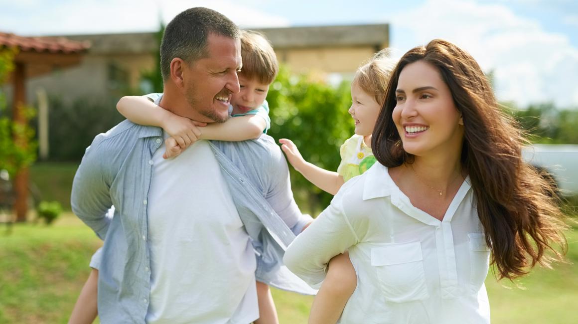 młodzi rodzice niosą na barana swoje dzieci, w tle dom na wsi