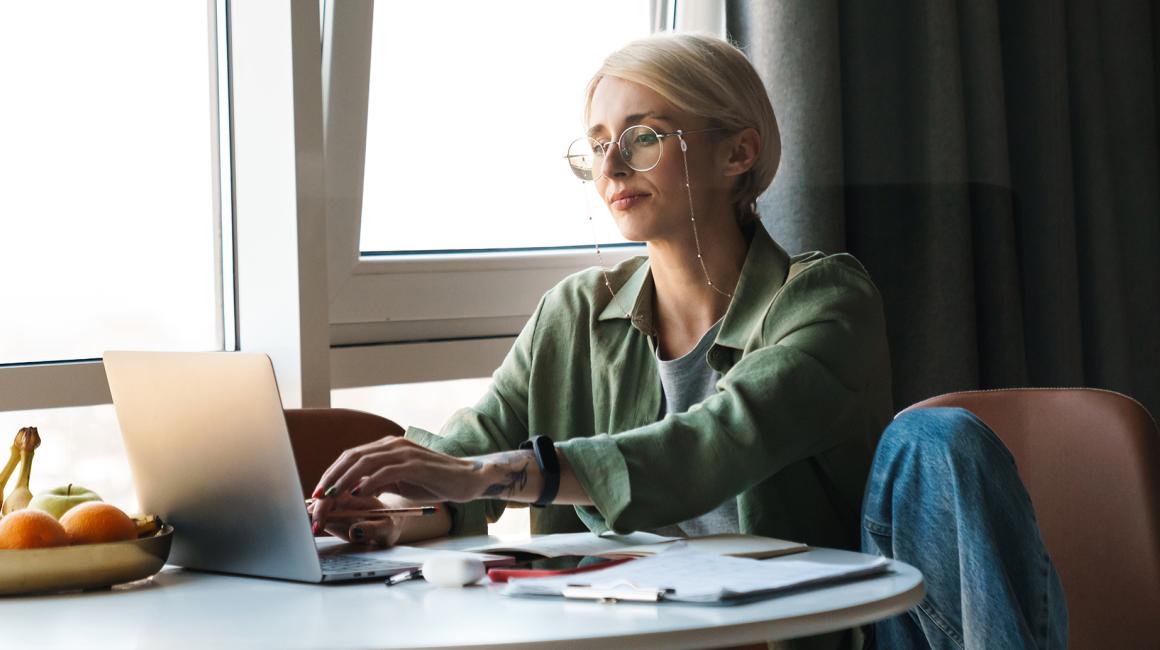 Blondynka w okularach przy laptopie leżącym na stoliku koło okna