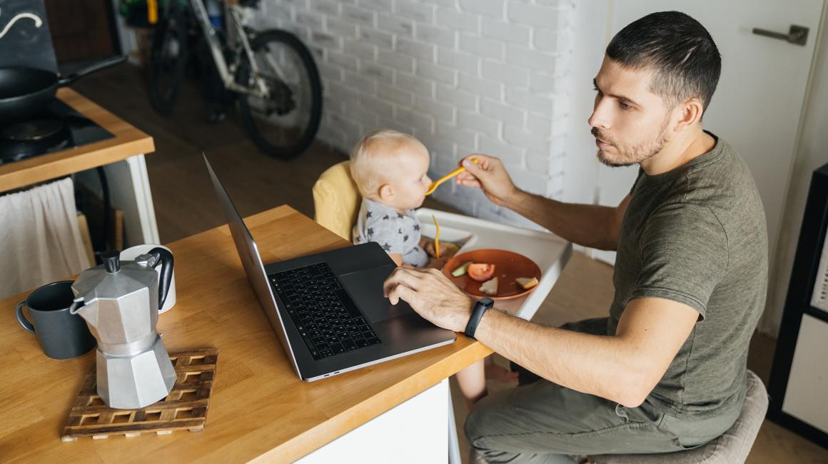 ojciec karmi dziecko łyżeczką i patrzy w laptop