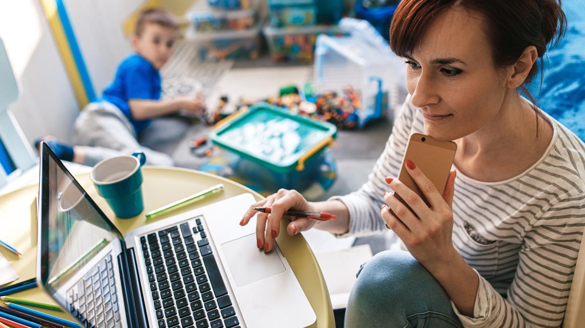 Mama przed komputerem, w jednej ręce komórka, w tle na podłodze bawi się chłopczyk