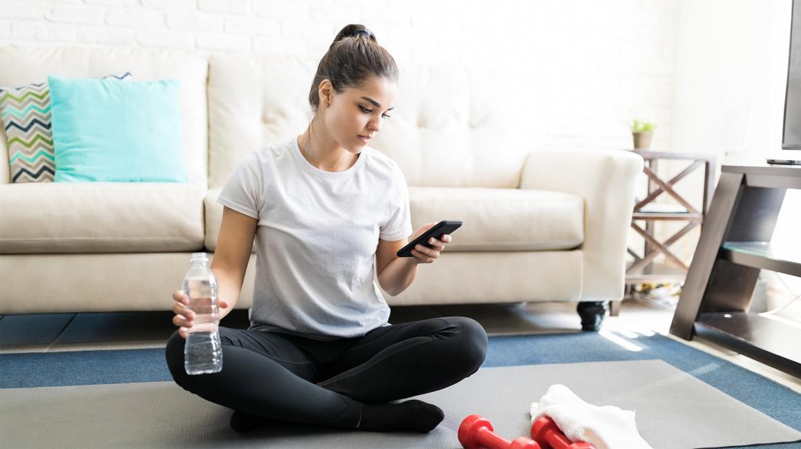 dziewczyna w bałym t-skircie siedzi po turecku w domu, ma w ręku butelkę wody, w drugiej komórkę